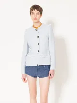 Chaqueta de estilo Chanel confeccionada en tejido jacquard, con cuello redondo, cierre frontal con botones y bolsillos delanteros con detalles de flecos. Presenta un corte entallado en la cintura con un ligero evasé en la parte inferior.