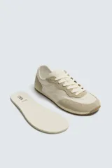 Zapatilla deportiva estilo barefoot, color marrón, con diseño de puntera ancha y redondeada. Confeccionada en una combinación de materiales textiles y detalles en serraje. Cuenta con suela fina de goma flexible y cordones en color crema.