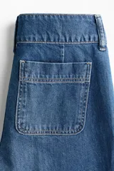 Jean de denim de algodón rígido, color azul, de tiro alto y pierna amplia con corte holgado. Presenta bolsillos delanteros insertados con detalles de ribete trenzado y pliegues marcados.