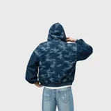 Campera canguro de denim con capucha, cierre frontal completo y bolsillos tipo parche, confeccionada en tela 100% algodón con diseño de camuflaje en tonos azules y detalles bordados.