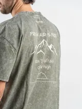 Remera de corte oversize color verde con efecto lavado, manga corta y cuello redondo. Presenta un estampado gráfico en la espalda con el diseño de una montaña y la frase 'Fresh air is free, don't be afraid of the high'.