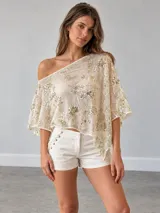 Top beige semitransparente con diseño de un solo hombro, mangas amplias y bordados metalizados con lentejuelas.