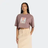 Remera cropped de manga corta, color marrón topo, con cuello redondo y un estampado frontal que muestra un par de zapatillas New Balance sobre un fondo de bloques de color degradados.