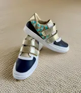 Championes de cuero con cierre de velcro, en combinación de colores blanco, negro y dorado texturizado, con detalles de arte pintado a mano en tonos turquesa y tostado en los laterales.