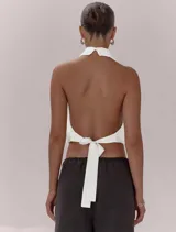 Top blanco con diseño halter, cuello alto y espalda descubierta. Presenta un frente cruzado con pliegues, bajo elástico y lazo de ajuste en la espalda.