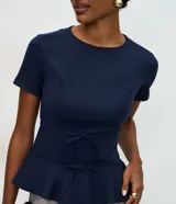 Blusa de punto roma color azul marino, con diseño estructurado, manga corta, recortes que definen la silueta, detalles de lazos decorativos en el frente y ruedo con volado peplum.