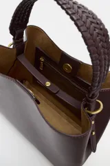 Bolso tipo saca de color bordó, con asa de mano trenzada y cierre mediante botón imantado. Incluye bolsillo interior con cremallera.