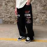 Pantalón de jean negro de corte super baggy con bordados de logos de marcas de automovilismo y texto en blanco y rojo.