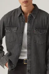 Camisa de jean estilo western en color gris lavado, con cuello clásico, cierre frontal con botones y dos bolsillos delanteros con solapa y botón. Presenta etiqueta roja distintiva en el bolsillo izquierdo.