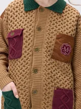 Campera tejido grueso y abrigado color verde militar, con forro de nylon interno, cuello estilo camisa en color negro y cierre frontal con botones. Cuenta con cuatro bolsillos delanteros con apliques en distintos tonos y texturas, y un parche bordado circular en el bolsillo superior izquierdo.