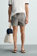 Bañador de hombre color verde claro con finas rayas blancas verticales (efecto seersucker). Tiene cintura elástica ajustable con cordón verde, bolsillos laterales y un bolsillo trasero de vivo.