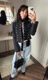 Chaqueta corta de denim negro con diseño de inspiración militar, cuello mao, cierre frontal con múltiples botones metálicos y detalles de trenzado horizontal. Presenta charreteras en los hombros y puños con botones decorativos.