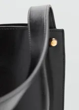 Bolso tipo saca de diseño minimalista en color negro, con asa de hombro fija y detalles de botones metálicos dorados en los laterales. Cuenta con cierre de mosquetón y forro interior.