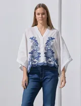 Blusa blanca con bordado floral azul, escote en V, manga japonesa y cierre frontal con botones.