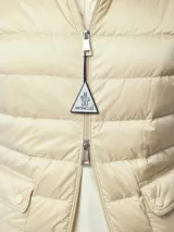 Campera corta acolchada Moncler modelo Lans, color beige, con cuello alto, cierre frontal con tirador personalizado, bolsillos laterales y logo de la marca en la manga izquierda.