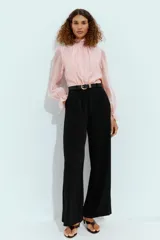Mono largo con parte superior de blusa rosa de manga larga, cuello alto fruncido y puños con volantes. La parte inferior es un pantalón negro de pernera ancha con bolsillos delanteros y un cinturón negro con hebilla dorada.