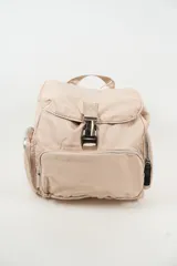Mochila de diseño casual en color beige, confeccionada en material sintético con acabado satinado. Presenta un compartimento principal con solapa y cierre de hebilla metálica a presión, además de bolsillos frontales y laterales con cierre de cremallera. Incluye asa superior y correas ajustables para los hombros.