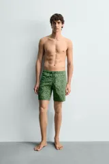 Bañador largo de tejido técnico color verde oscuro con estampado de flores en tono verde más claro. Presenta cintura elástica ajustable con cordón, bolsillos laterales y un bolsillo trasero de plastrón.