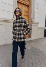 Campera de algodón con estampado de cuadros tartán en tonos negro y beige. Presenta cuello alto con cierre de botones metálicos, mangas largas y un cinturón ajustable en la cintura para entallar la silueta.