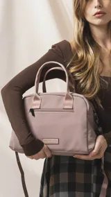Cartera de mano color beige, confeccionada en tela impermeable, con doble asa corta y correa larga ajustable y desmontable. Presenta cierre principal, bolsillo frontal con cierre y detalle de colgante decorativo.