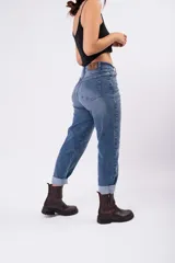 Pantalón tipo mom jean de tiro alto, confeccionado en denim rígido de color azul medio con efecto lavado. Presenta un corte recto y holgado con ruedos doblados, cierre frontal con botón y bolsillos laterales.