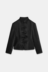 Chaqueta negra de tejido satinado, con cuello mao, manga larga con hombreras, corte evasé y cierre frontal con alamares.