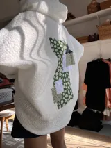 Campera tipo hoodie de corderito blanco con diseño de parches en la espalda que combinan telas estampadas con motivos florales en tonos verdes y detalles de encaje blanco.