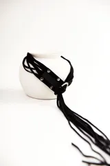 Cinturón negro de cuero con hebilla metálica plateada en forma de aro. El cinturón se anuda y tiene una tira de gamuza negra con flecos colgantes.