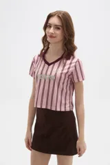 Remera estilo jersey de fútbol con corte cropped, cuello en V y mangas cortas. Presenta un diseño de rayas verticales en tonos rosados y bordó, con estampado frontal que incluye el texto 'TOKYO' y el número '24'.