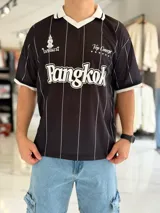 Camiseta deportiva negra de manga corta con diseño estilo jersey de fútbol, rayas verticales blancas y cuello polo blanco con detalles negros. Presenta estampados en el frente, incluyendo la palabra 'Bangkok' en gran tamaño.