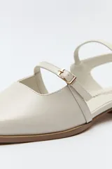 Zueco destalonado de charol color crema, con punta cuadrada, taco bajo y dos tiras finas con hebillas metálicas ajustables en el empeine y el talón.