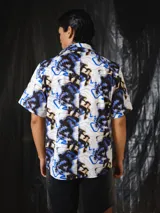 Camisa de manga corta de calce relajado, con cuello clásico, cierre frontal con botones y bolsillos aplicados en el pecho. Presenta un estampado integral abstracto en tonos crudo, negro, marrón y azul eléctrico.