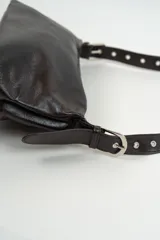 Cartera de hombro color marrón oscuro, confeccionada en cuero sintético con acabado brillante. Presenta una correa larga ajustable con detalles de piezas circulares decorativas y remaches metálicos.