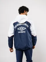 Campera de lluvia Umbro del Club Nacional de Football, con diseño de bloques de color en rojo y azul marino. Presenta capucha, cierre frontal, puños elásticos y detalles de piping blanco. Incluye logo de Umbro y escudo del club en el pecho.