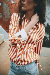 Camisa de manga larga con estampado de rayas verticales en color naranja y crema. Presenta cuello solapa, cierre frontal con botones dorados y puños anchos en color crema con detalle de lazos ajustables.
