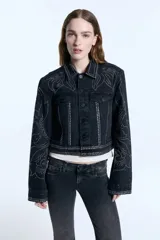 Campera de denim negro con corte estructurado a la cintura, decorada con patrones de tachas metálicas en hombros, mangas y frente. Presenta cierre frontal con botones, bolsillos con solapa y puños abotonados.