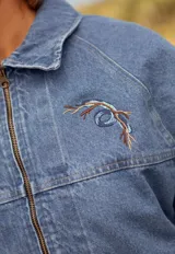 Campera de jean con mangas y frente en denim azul, y panel trasero estampado con motivos musicales y florales en tonos tierra. Incluye un parche de cuero con el logo de la marca en la parte superior de la espalda.