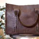 Cartera de cuero color tostado, estructurada, con doble asa de mano, correa desmontable y dos bolsillos frontales verticales con cierre. Presenta detalles de costuras pespunteadas y el logo de la marca grabado en la parte inferior.