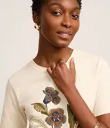 Remera de algodón color beige con cuello redondo y mangas cortas. Presenta un estampado frontal con diseño floral de estilo mosaico y texto en la parte inferior.
