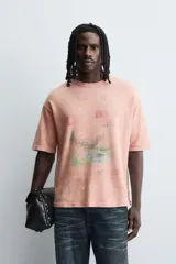 Camiseta de punto de algodón con corte boxy fit, cuello redondo y manga corta. Presenta un estampado frontal con diseño de paisaje y texto en tonos deslavados. La prenda tiene un efecto lavado que le da un aspecto envejecido e irregular.