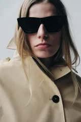 Trench corto color verde militar, con cuello solapa y capucha. Tiene manga larga con trabillas y botón en los puños, bolsillos delanteros y bajo asimétrico. Cierre frontal con botones. El tejido es repelente al agua.