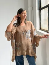 Blusa holgada color camel con estampado floral en tonos grises y azules, cuello redondo y mangas amplias con volados, al igual que el ruedo inferior.