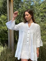 Vestido corto blanco de lino con cuello mao y cierre parcial de botones de coco. Presenta un diseño amplio con varios volados en el cuerpo y mangas acampanadas de tres cuartos.