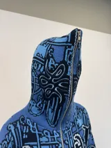 Buzo tipo hoodie tejido de punto grueso con cierre frontal completo hasta la capucha. Presenta un diseño gráfico abstracto en tonos azules y negros, con tipografía estilo gótico en el frente y las mangas. Cuenta con puños y ruedo acanalados en color negro, y etiqueta de marca en la manga.