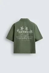 Camisa de corte relajado color verde oliva, con cuello solapa, manga corta y cierre frontal de botones. Presenta un estampado con el texto 'ASTRAL Drift' en el frente.