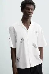 Camisa de manga corta confeccionada en mezcla de algodón y lino, color blanco. Presenta un cuello tipo bowling, calce regular y cierre frontal con botones. Destaca por sus bordados lineales en contraste de color negro con motivos de frutas y hojas distribuidos por toda la prenda.
