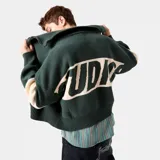 Campera de tejido de punto color verde oscuro con cuello tipo polo y cierre frontal. Presenta franjas anchas color beige en el pecho con la inscripción "EME" en letras grandes y franjas a juego en las mangas. Corte oversize.