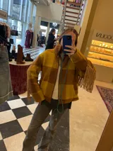 Campera de lana italiana con diseño de cuadros en tonos amarillo y marrón. Presenta flecos de gamuza en las mangas, cierre frontal y bolsillos laterales. La prenda se encuentra completamente forrada.