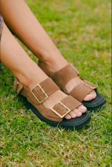 Sandalia de estilo birkenstock color marrón, con doble tira y hebillas metálicas ajustables, y suela plana.