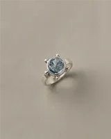 Anillo de plata 925 con piedra central redonda facetada de topacio celeste, con diseño de banda chata y detalles de pequeñas esferas alrededor de la piedra.